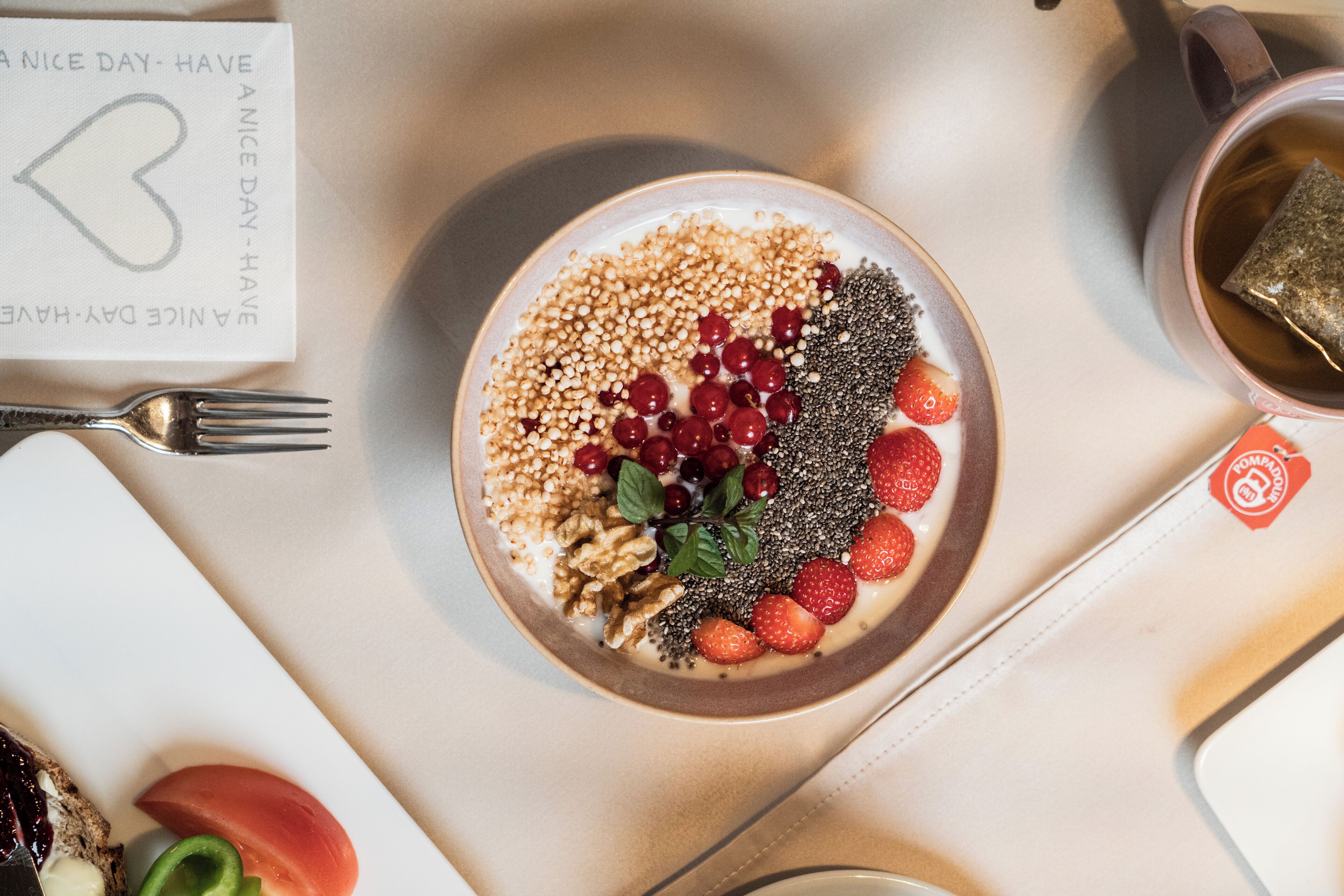 Een ontbijttafel met een kom yoghurt belegd met granola, bessen en zaden, een kopje thee, en een kaartje met een hartsymbool.