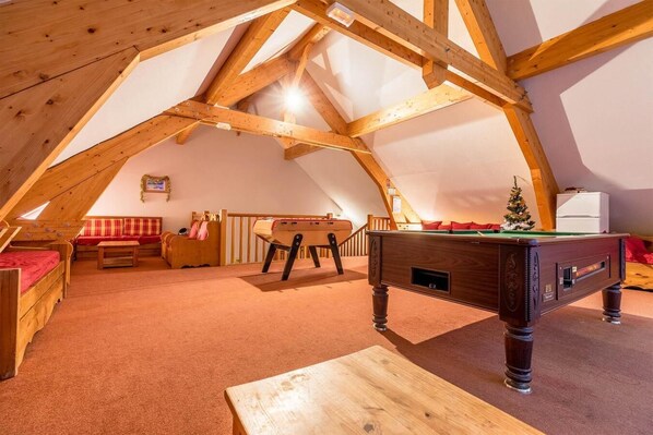 Une pièce mansardée chaleureuse avec un plafond en poutres apparentes, une table de billard, un canapé et un petit sapin de Noël.