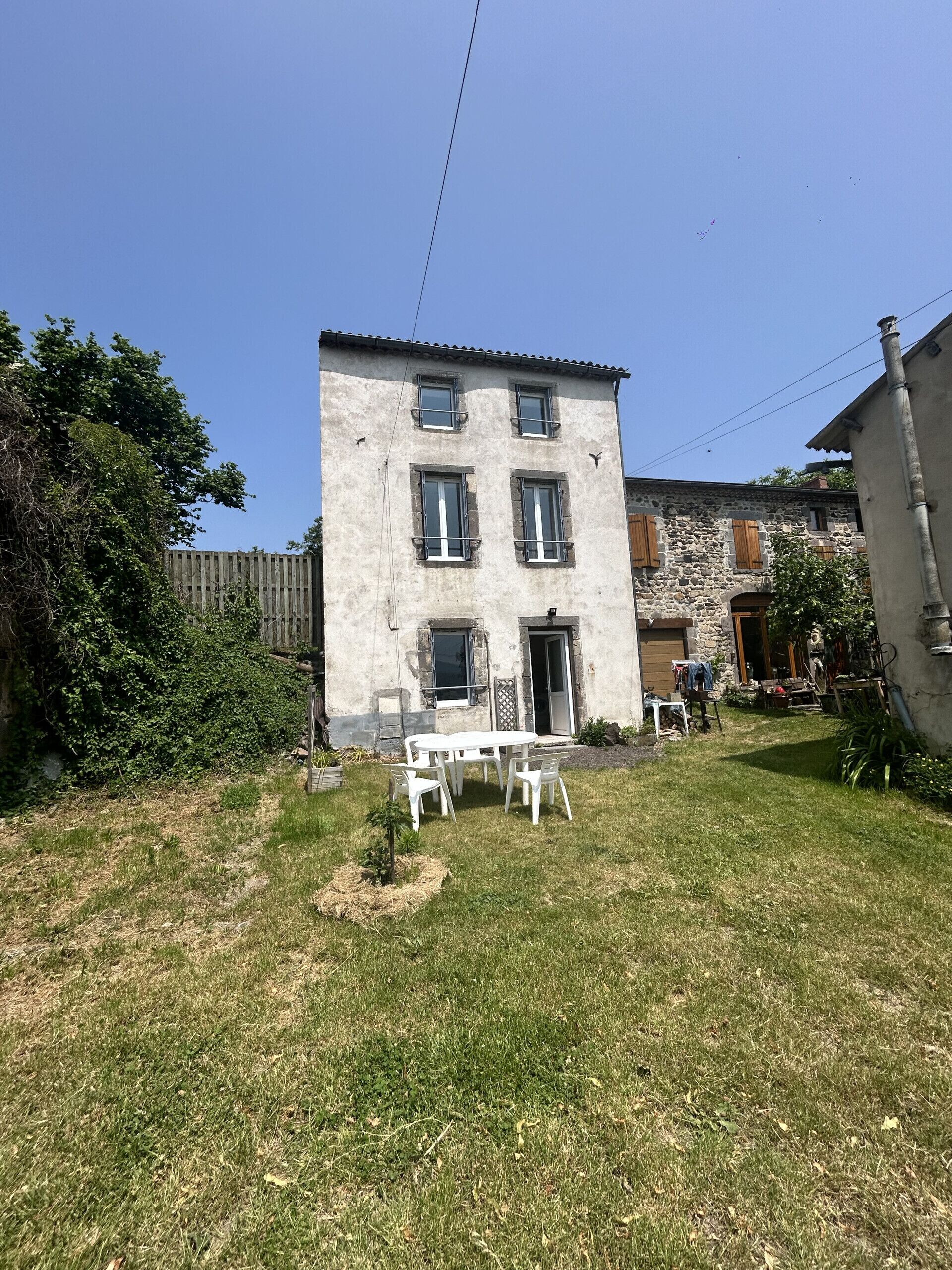 A two-story building with a stone facade, a small front yard with a white table and chairs, and a wooden fence.