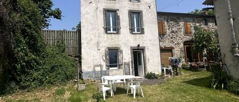 A two-story building with a stone facade, a small front yard with a white table and chairs, and a wooden fence.
