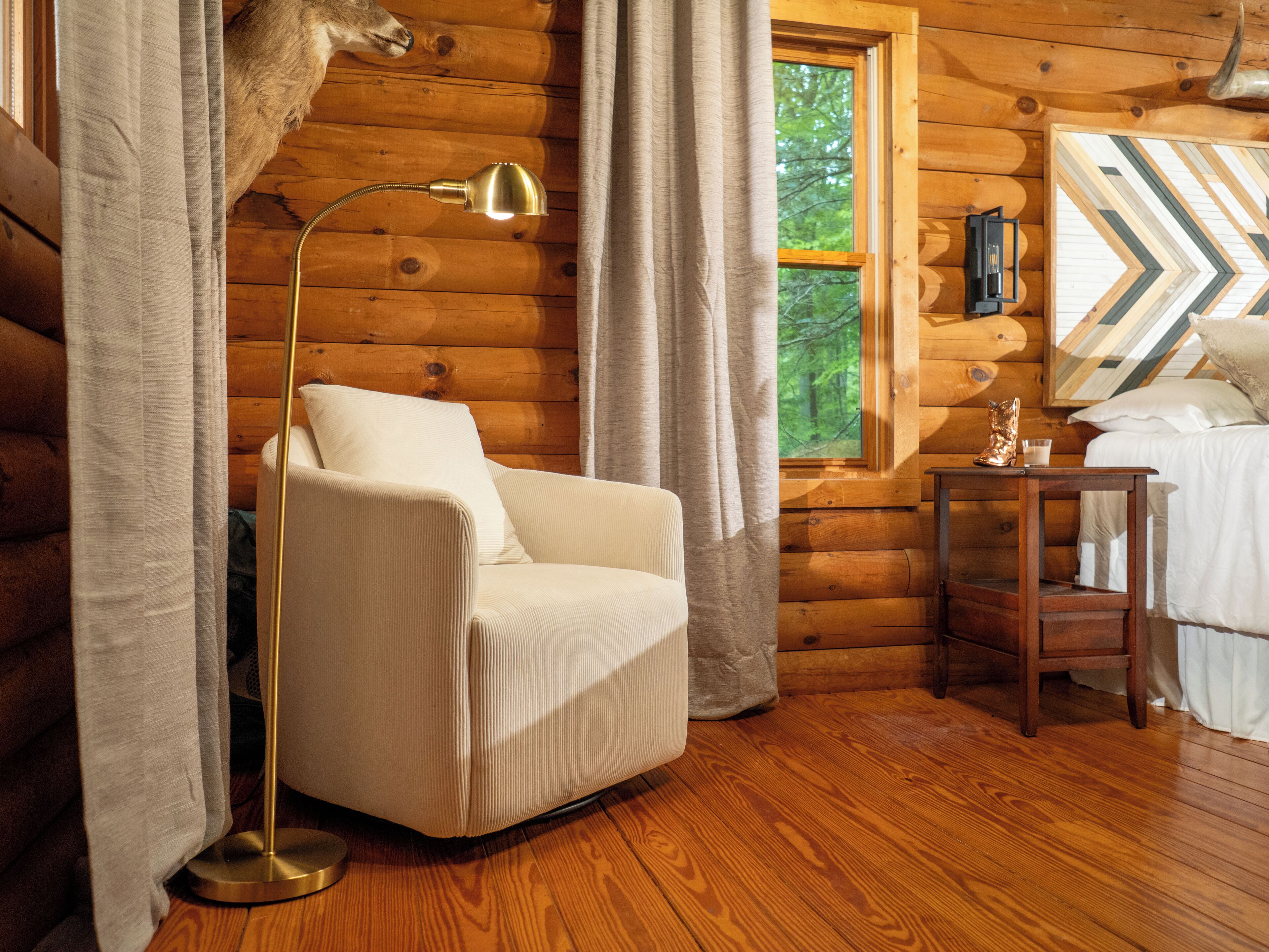 Una habitación tipo cabaña de madera con un sillón blanco, una lámpara de pie, una mesa de madera y una cama con cabecera estampada en patrón zigzag.
