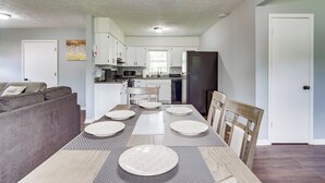Een moderne keuken met witte kastjes, een eettafel gedekt met placemats, en een grijze bank in de woonkamer.