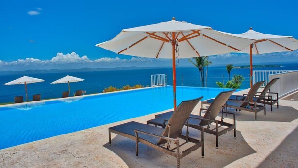 Un espace aménagé au bord de la piscine, avec des chaises longues et des parasols, offrant une vue sur un océan d’un bleu limpide et sur des montagnes lointaines.