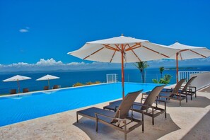 A poolside area with lounge chairs and umbrellas, overlooking a clear blue ocean and distant mountains.