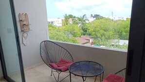 A balcony with two chairs and a small table, a dreamcatcher, and a view of buildings and trees. - Studio in Centro Maya area of Playa Del Carmen-full kitchen, AC, smart TV, pool. (Playa del Carmen)