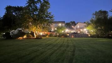 Um imóvel residencial bem iluminado ao anoitecer, com um gramado extenso, um caminho de pedras que leva à entrada e uma casa de vários andares com janelas visíveis e uma varanda.