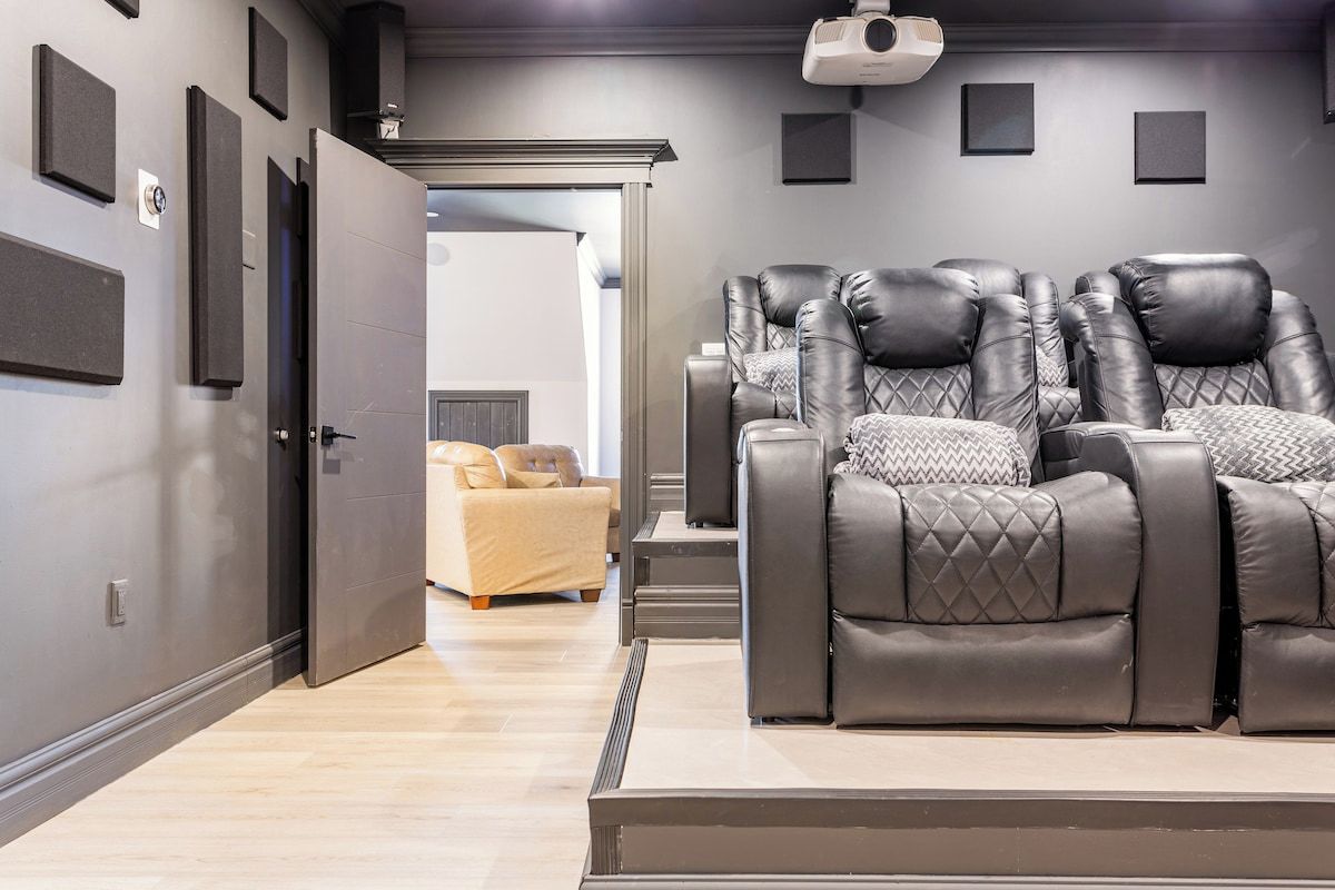 Una sala de cine en casa con un sofá de cuero reclinable, un sofá color beige y un proyector montado en el techo.