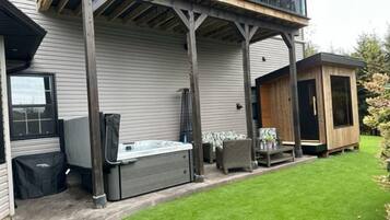 Een huis met een overdekt terras, een jacuzzi en een klein houten bijgebouw.