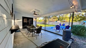 Un patio techado con mesa y sillas, una parrilla de barbacoa y vistas a un jardín trasero con piscina y sillas.