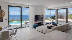 Een moderne woonkamer met een grote televisie, een eethoek met een gedekte tafel, en uitzicht op zee.
