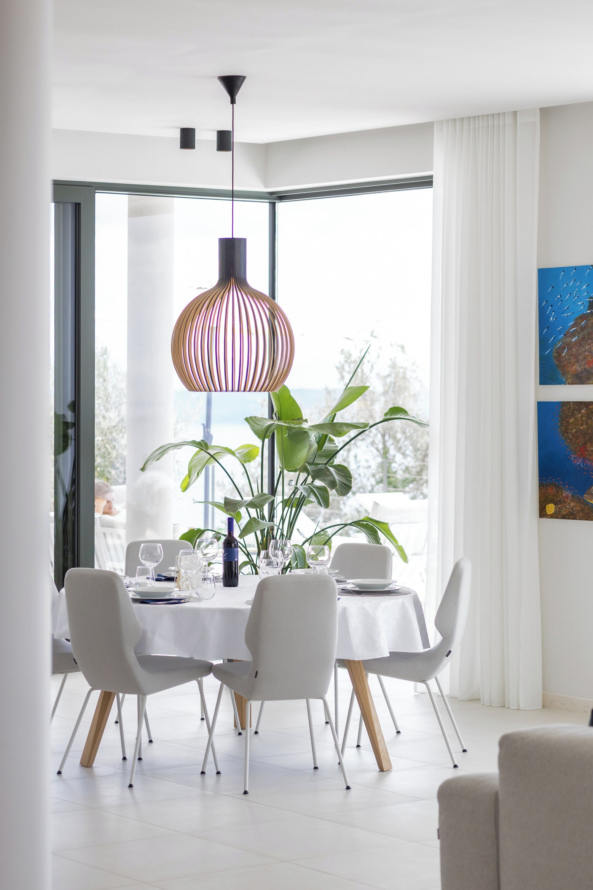 A modern dining area with a white table, white chairs, a striped pendant light, and a large window with curtains.