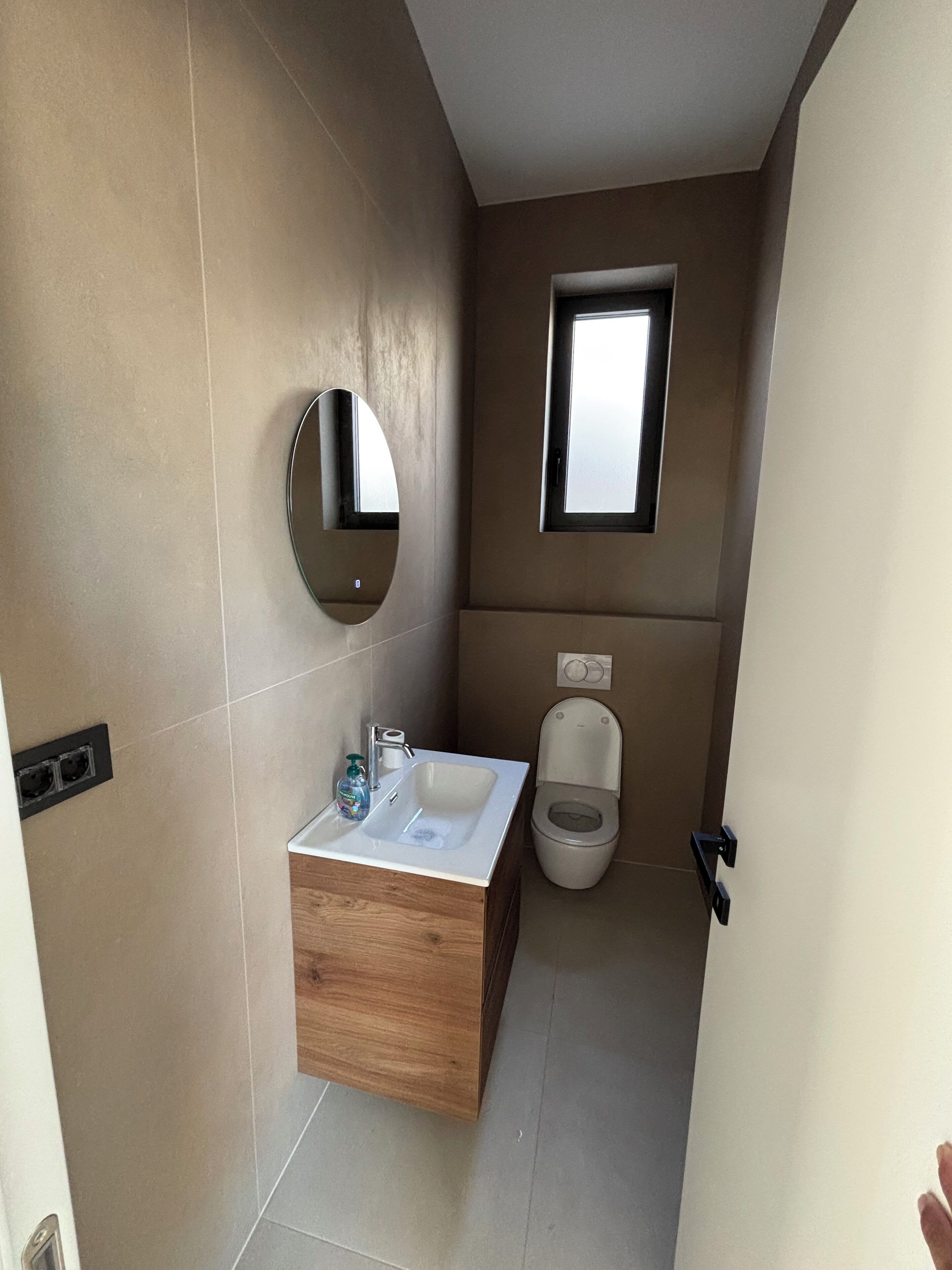 Un bagno compatto con un mobiletto in legno, uno specchio rotondo, una piccola finestra e un water.