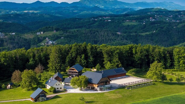 A large, modern building with multiple sections, surrounded by lush green fields and forested hills. - Posestvo Pule Equestrian Estate (Mokronog-Trebelno)