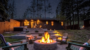 A circular fire pit surrounded by Adirondack chairs, a wooden deck, and a house with a porch.