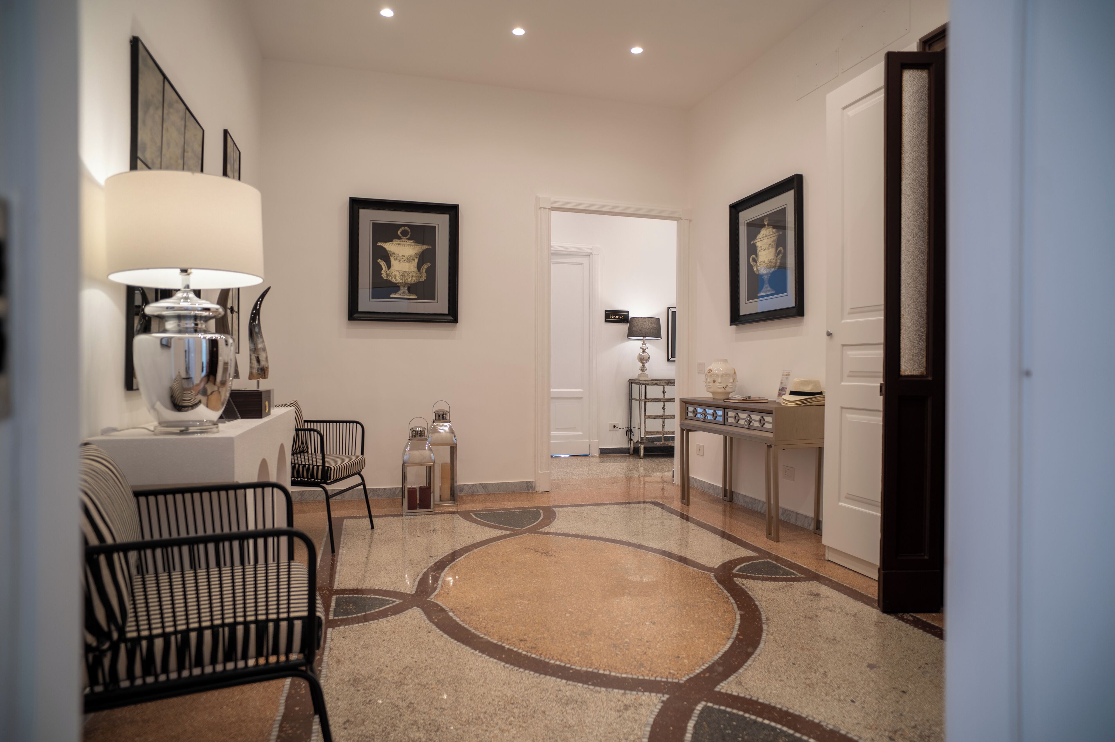 A hallway with a patterned floor, a lamp, framed artwork, and a door leading to another room.