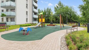 Ein Spielplatz mit Rutsche, Schaukeln und einer Bank, umgeben von einem befestigten Bereich und Grünflächen.