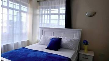 Un dormitorio con cama, cortinas blancas, una mesita de noche y un jarrón con flores.