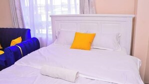 Une chambre d’hôtel avec un lit blanc, un fauteuil bleu et une fenêtre avec des rideaux.