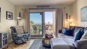 Un salon comprenant un canapé, un fauteuil et une table agrémentée d’objets décoratifs. Une porte-fenêtre coulissante donne accès à un espace extérieur avec des chaises et une vue sur l’océan.