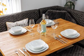 Una mesa de comedor dispuesta con platos blancos, vasos, cubiertos, una botella de vino y un cojín decorativo con la inscripción "HAPPINESS IS".