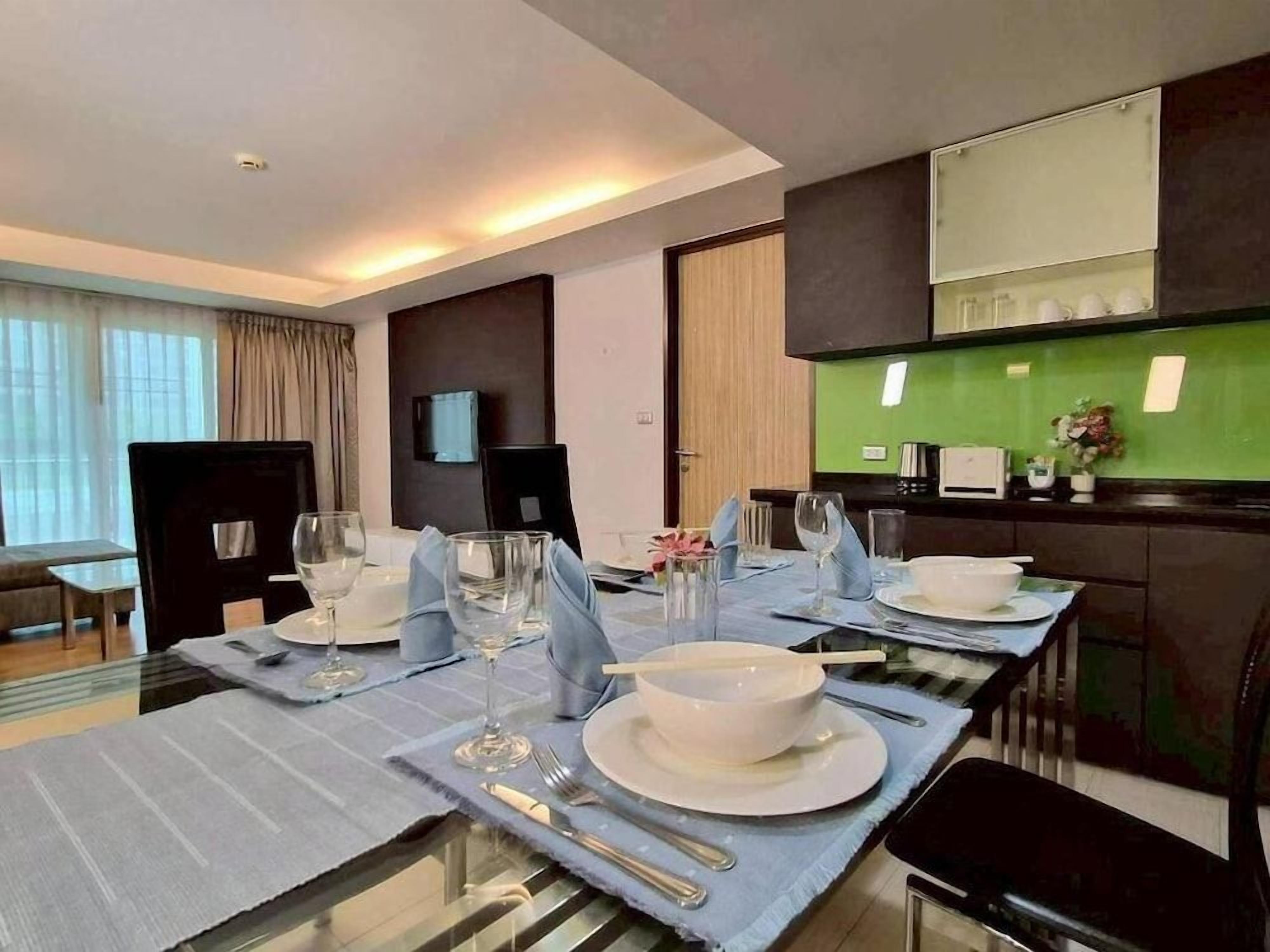 A modern hotel dining area with a long table set for a meal, a television mounted on the wall, and a kitchen area visible in the background.