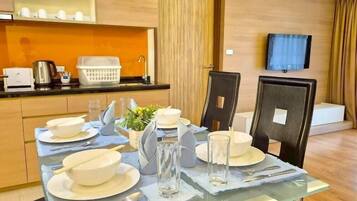 A modern dining area with a glass table set for four, including bowls, plates, and cutlery. A coffee maker and a kettle are on the counter, and a television is mounted on the wall.