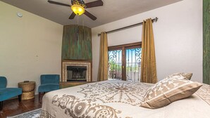 A bedroom with a large bed, a ceiling fan, a fireplace, and a view through a window with curtains.