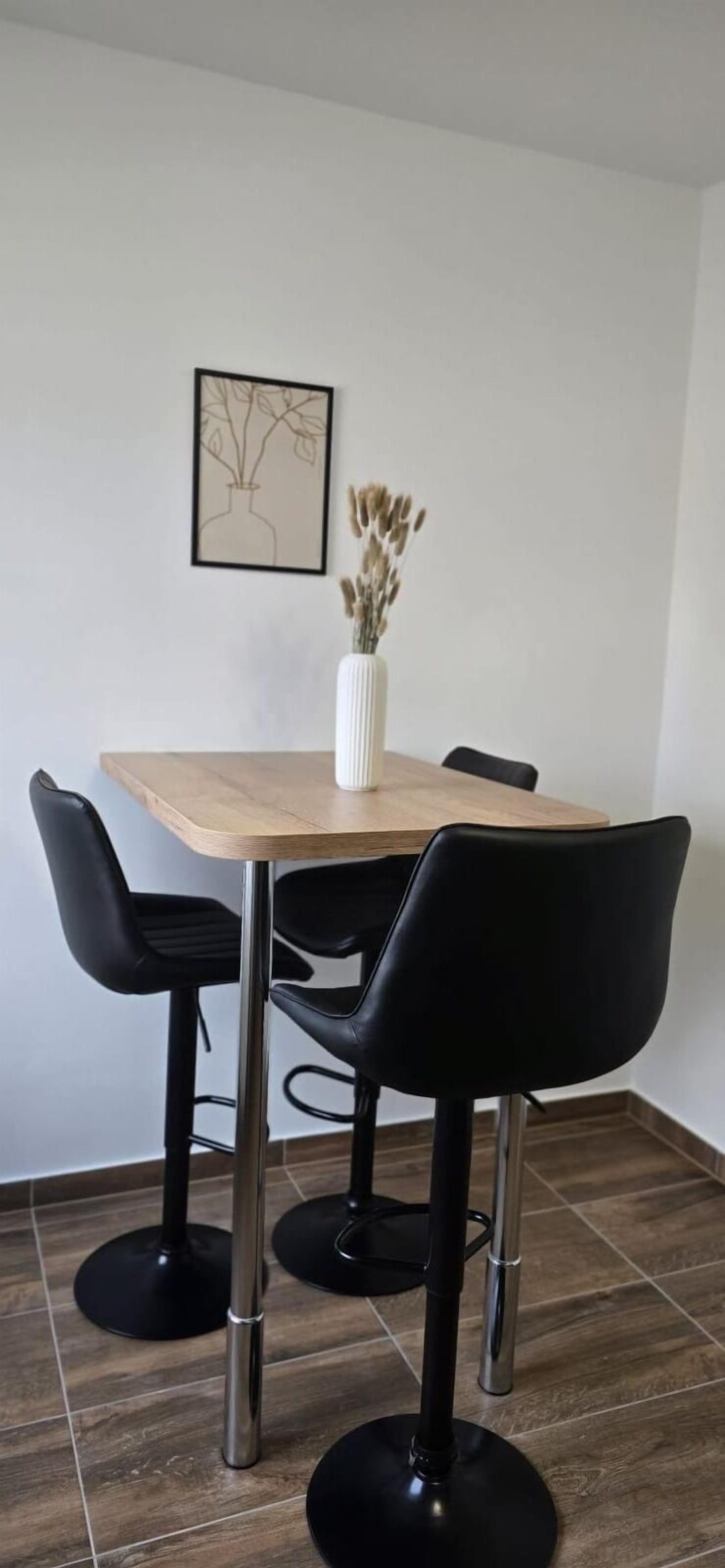Ein Essbereich mit einem Holztisch, vier schwarzen Stühlen, einer weißen Vase mit Trockenpflanzen und einem gerahmten Bild an der Wand.