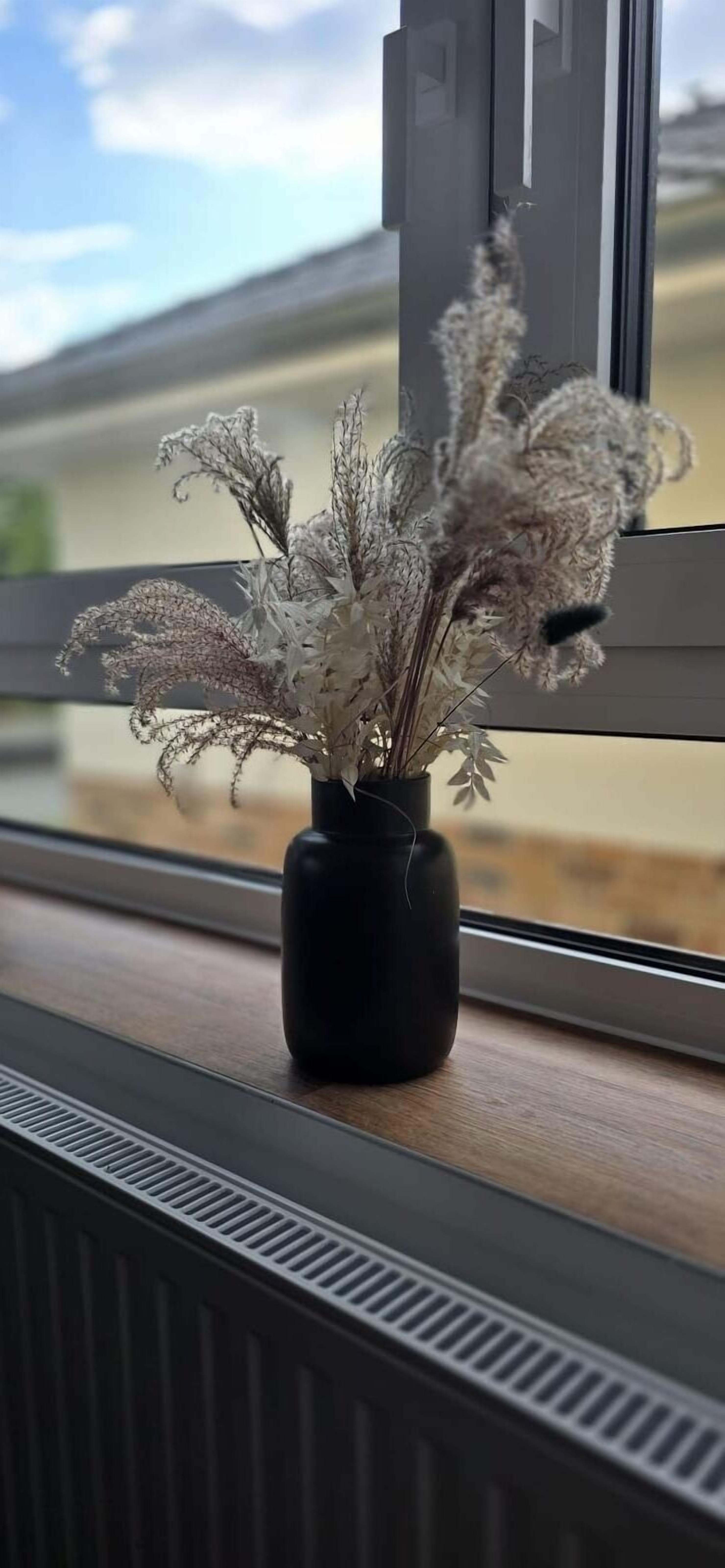 Eine schwarze Vase mit getrockneten Blumen auf einem Fensterbrett.