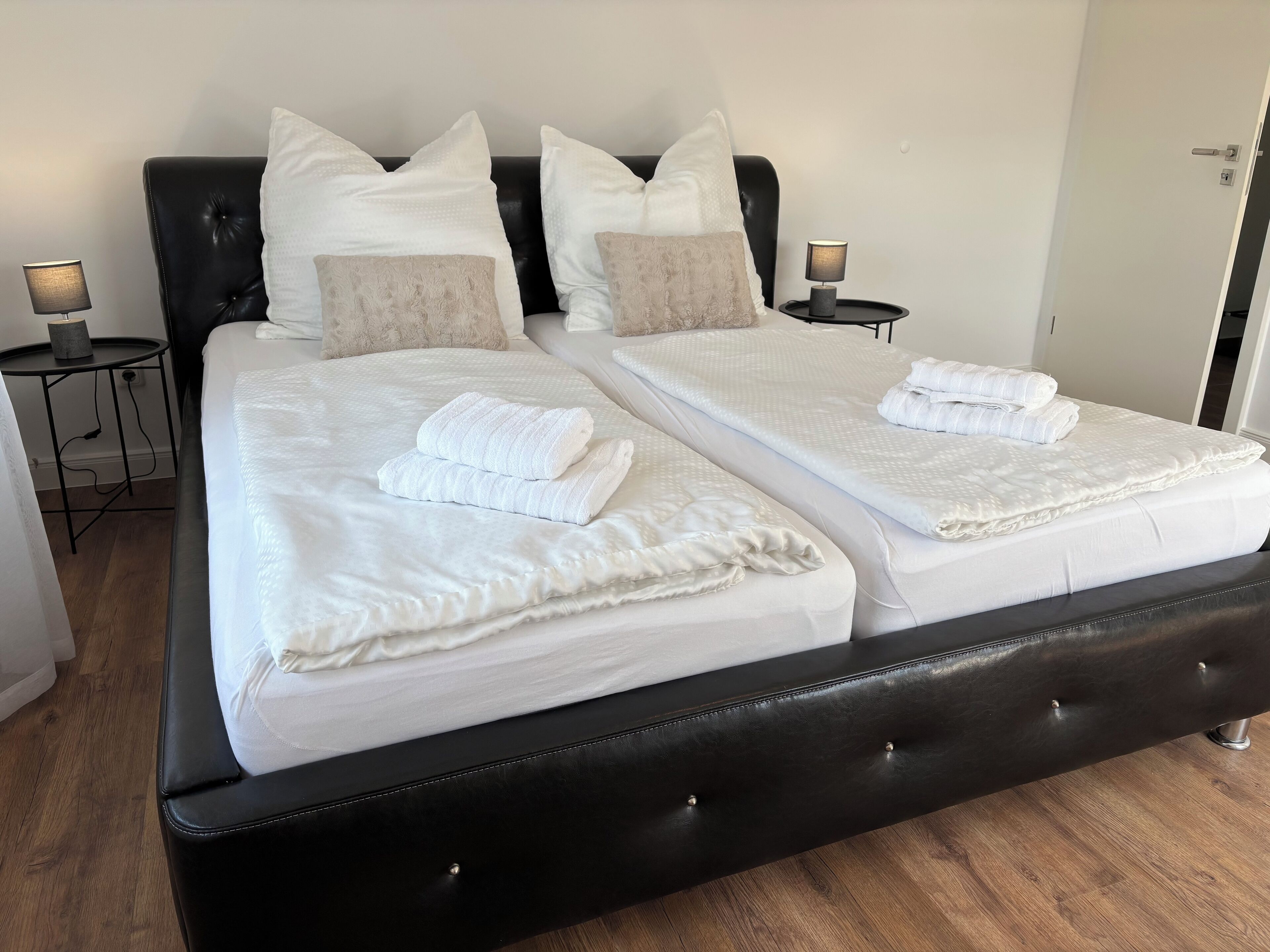 Uma cama de casal com lençóis brancos e duas almofadas, cabeceira preta e duas mesas de cabeceira com abajures.
