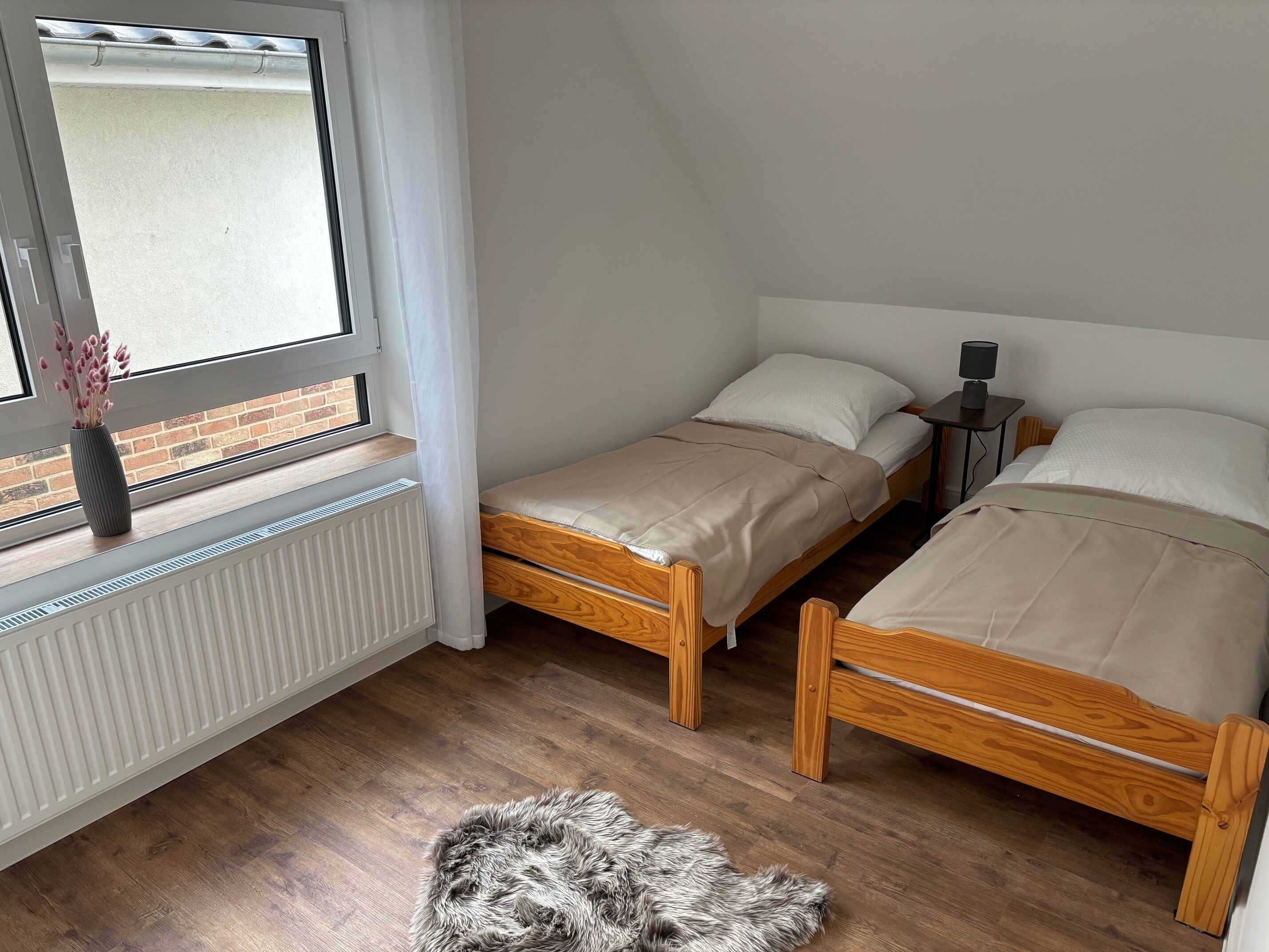 Un dormitorio con dos camas de madera, un radiador, una ventana que da a un muro de ladrillo y una pequeña mesa con una lámpara.