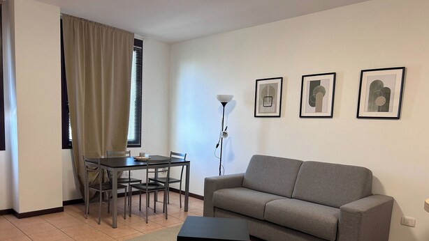 A living room with a grey sofa, a small black table, a dining table with chairs, and three framed pictures on the wall. - Apartment 3 B (Emilia-Romagna)