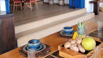 Zona pranzo con tavolo di legno apparecchiato per due persone, una sedia blu e un centro tavola con frutta tropicale.