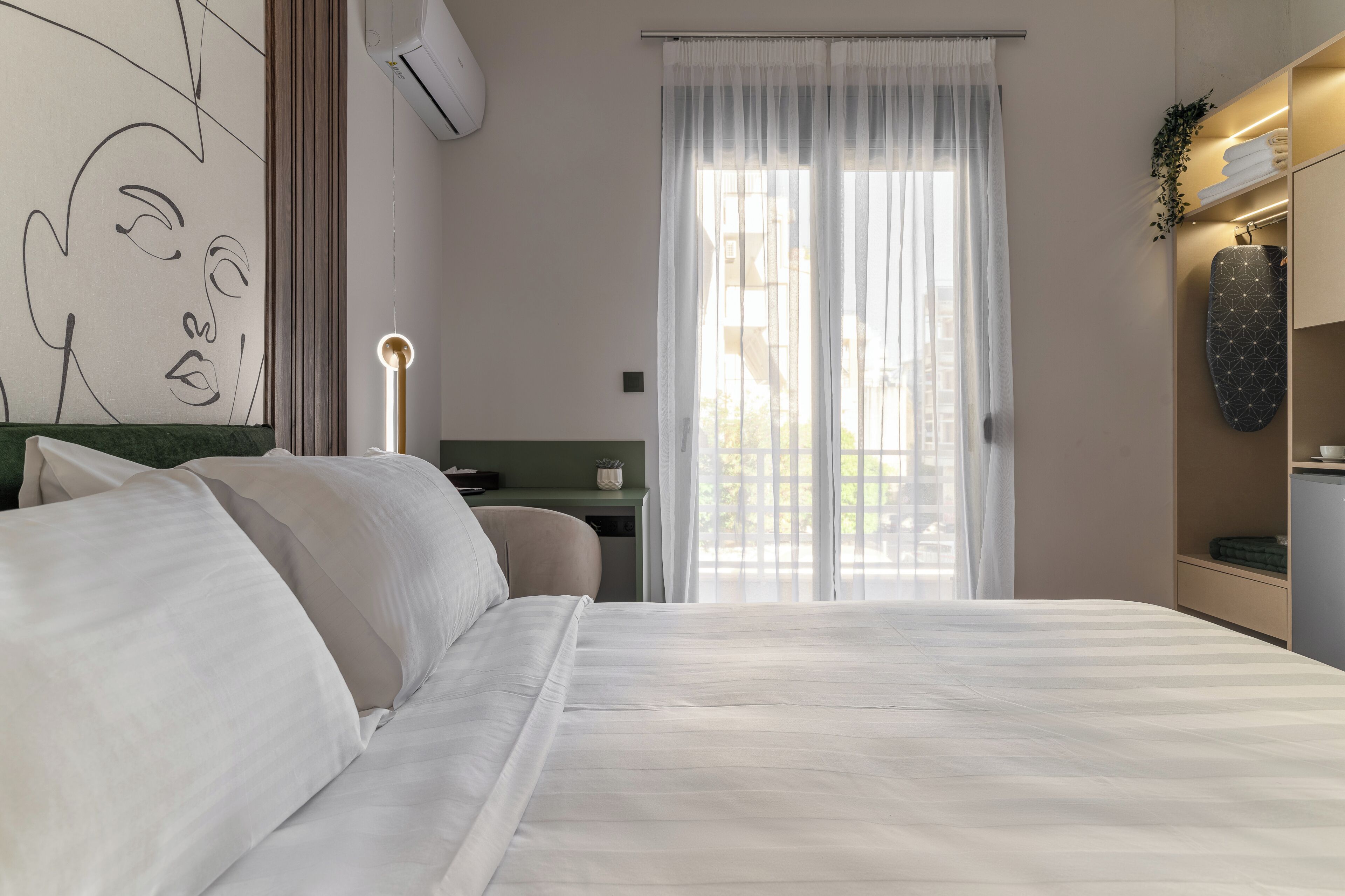 A bedroom with a large bed, a desk, a chair, a wall-mounted air conditioner, and a window with sheer curtains.