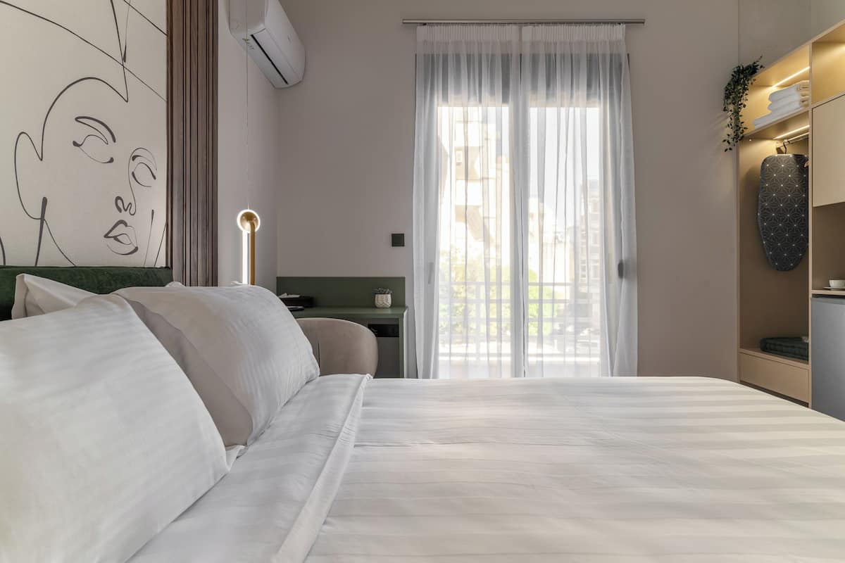 A bedroom with a large bed, a desk, a chair, a wall-mounted air conditioner, and a window with sheer curtains.