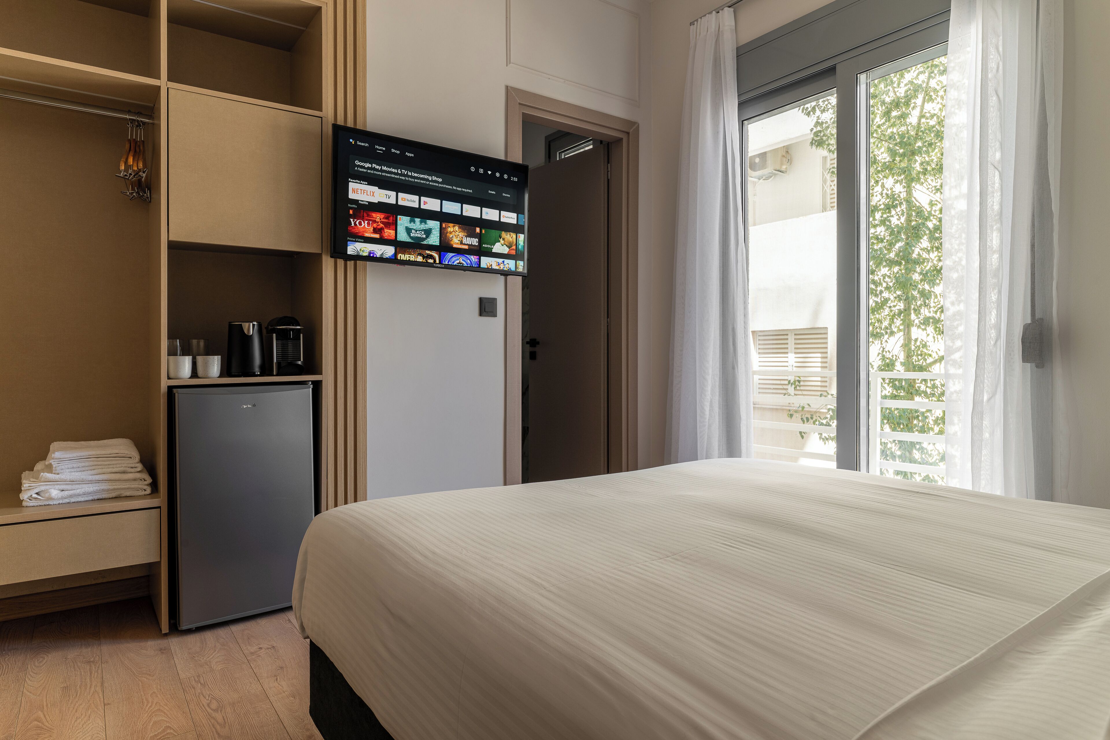 Ein Hotelzimmer mit einem Bett, einem an der Wand befestigten Fernseher, der einen Streaming-Dienst zeigt, einer kleinen Kühlbox und einem Kleiderschrank mit darin befindlichen Kleidungsstücken.