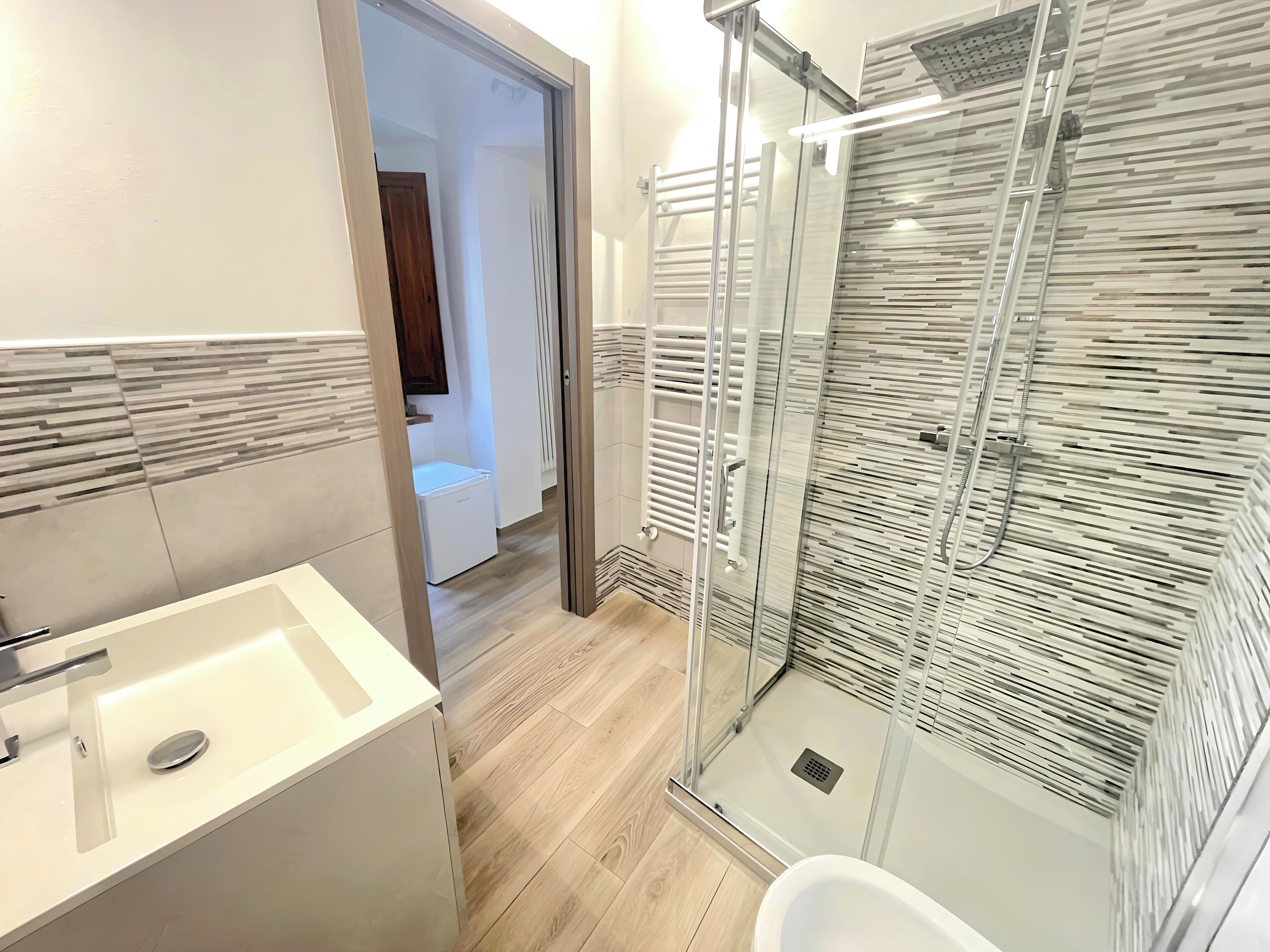 A modern bathroom with a glass shower enclosure, a white sink, and a wooden floor.