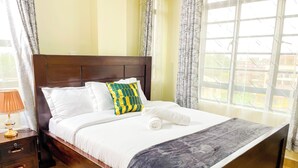 A neatly made bed with white bedding and a patterned pillow, a wooden headboard, a nightstand with a lamp, and a view through large windows.