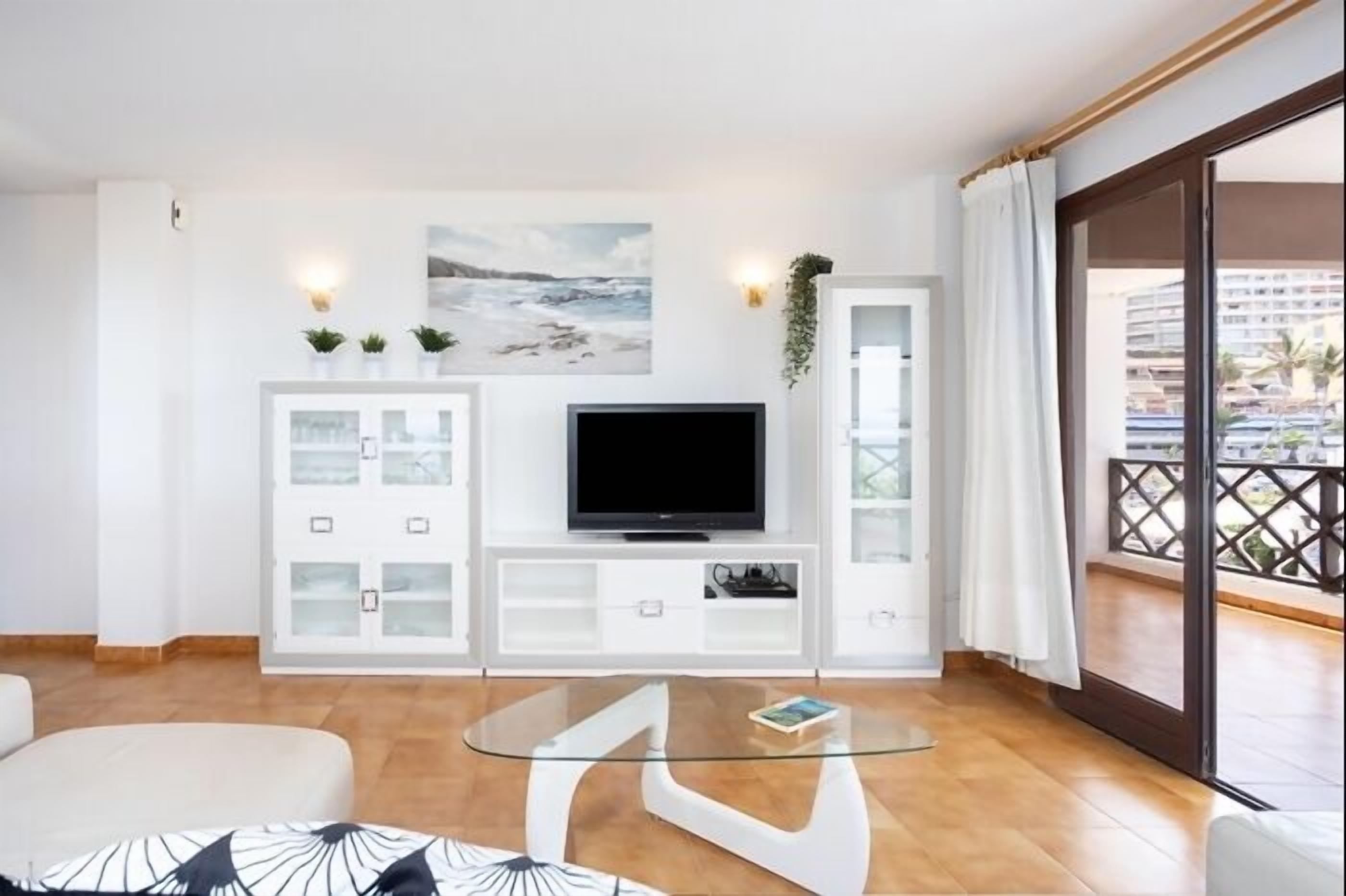 A modern living room with a glass coffee table, a flat-screen TV, and a balcony with a view of the city.