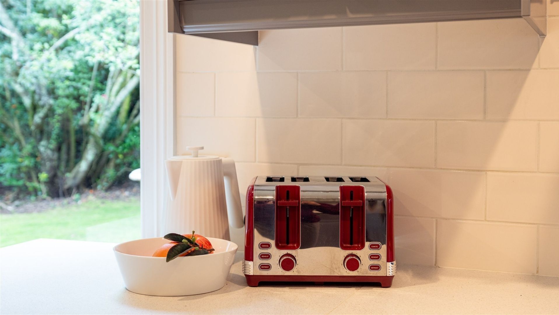 Een keuken met een rode toaster, een witte schaal met fruit en een witte karaf.