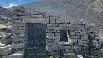 A stone structure with an open doorway, surrounded by rocks and grass, set against a mountainous backdrop.