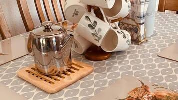 Un coin cuisine avec une bouilloire en acier inoxydable posée sur un sous-verre en bois, une pile de tasses blanches ornées de feuilles vertes et une boîte à biscuits.