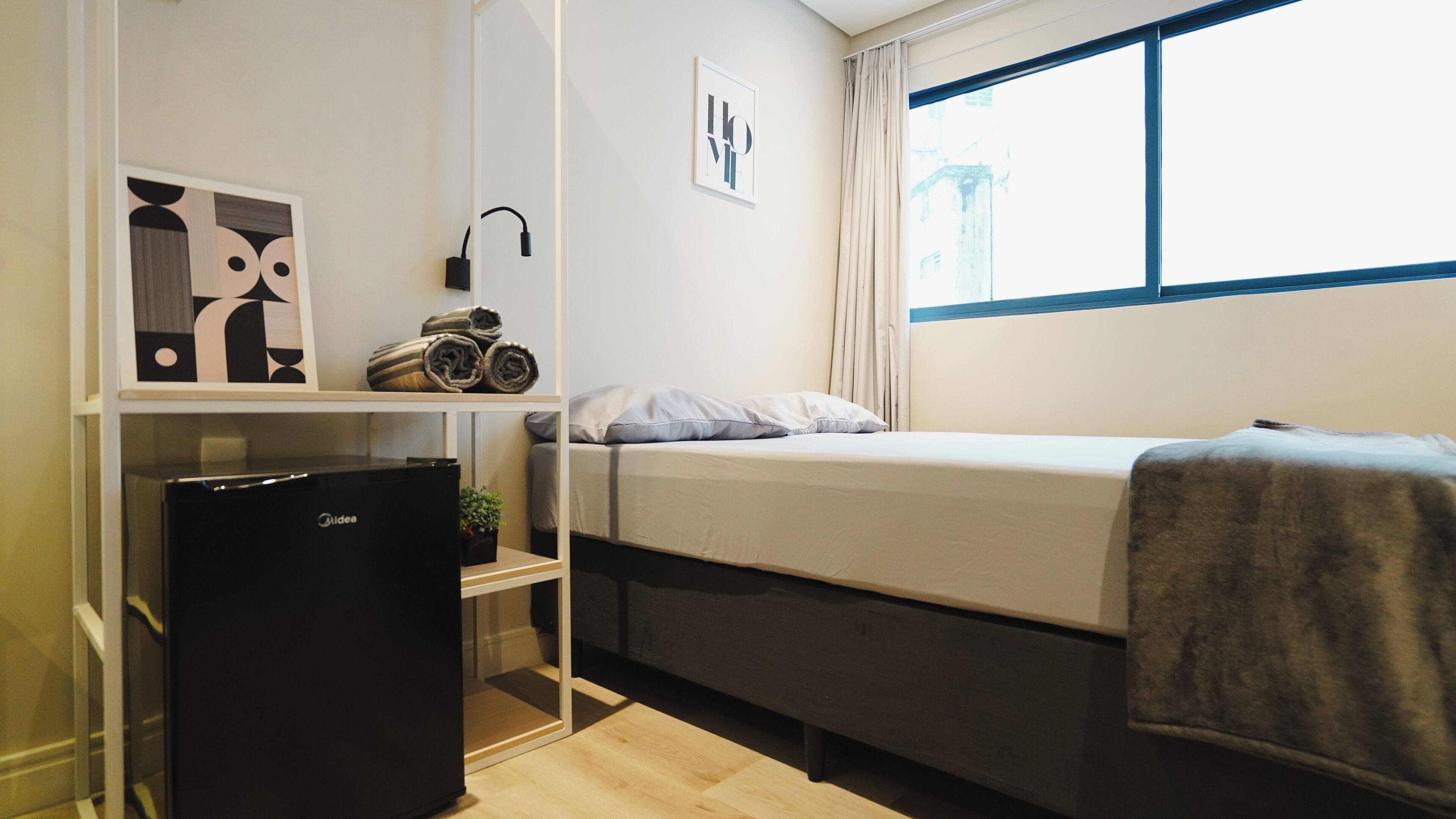 A compact room with a bed, a small desk with a computer, a refrigerator, and a window with curtains.