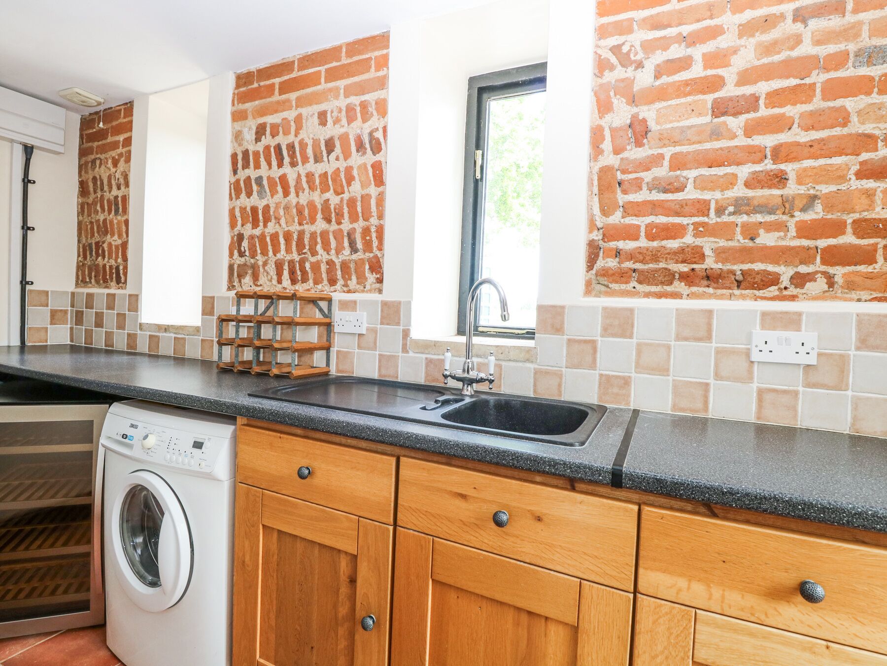 Una cocina con lavadora, armarios de madera, encimera negra, lavabo y ventana con vistas al verdor.