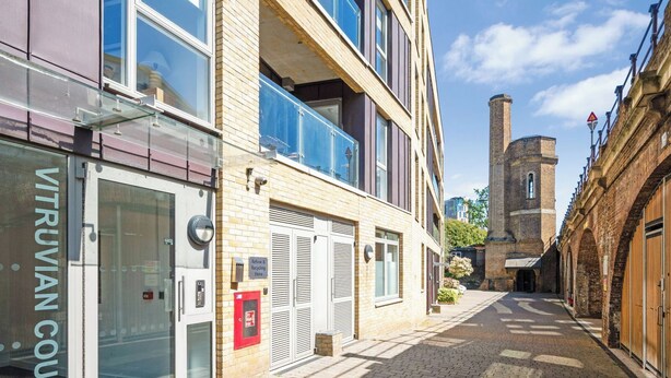 Une rue étroite bordée d’un bâtiment moderne dont la façade est en verre et sur laquelle on peut lire l’inscription « VITRUVIAN COURT », avec une tour de briques traditionnelle en arrière-plan.