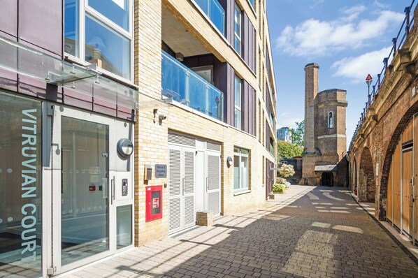 Une rue étroite bordée d’un bâtiment moderne dont la façade est en verre et sur laquelle on peut lire l’inscription « VITRUVIAN COURT », avec une tour de briques traditionnelle en arrière-plan.