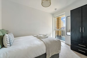 A bedroom with a bed, pillows, a blanket, a wardrobe, and a view through a window. - Canary Exquisite - 3 Bedroom Apartment (Greater London)