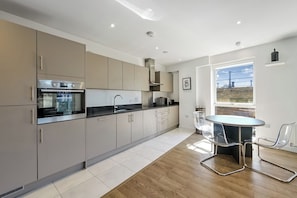 A modern kitchen with built-in appliances, a dining area with a round table and chairs, and a view through a window. - Canary Exquisite - 3 Bedroom Apartment (Greater London)