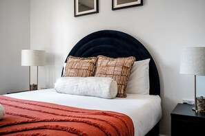A neatly made bed with a dark headboard, white bedding, and patterned pillows. There are two bedside tables with lamps, and the room has a minimalist decor.