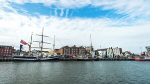 Un puerto con varios barcos de vela históricos amarrados a lo largo del muelle, rodeado de edificios con comercios.